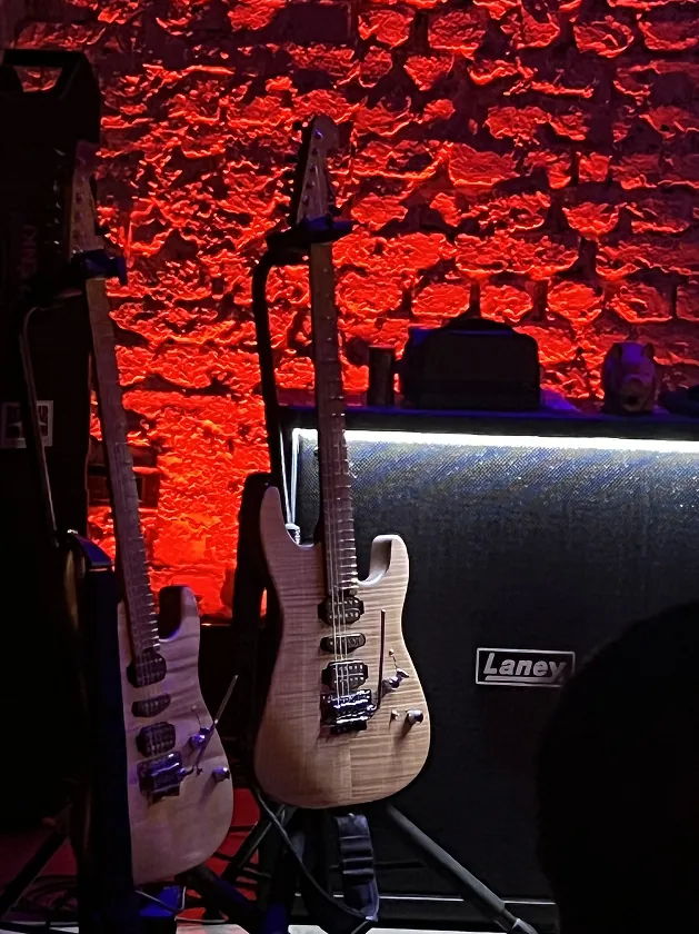 Two electric guitars on stands in front of a red-lit brick wall.